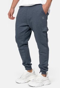 Mężczyzna w ciemnoszarych dresowych spodniach z boczną kieszenią i białych, masywnych sneakerach, stojący na jednolitym tle.