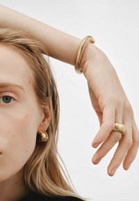 La joyería en tono dorado incluye una pulsera de asta texturizada, un anillo a juego y pendientes en espiral, que exhiben un diseño moderno y elegante con un brillo metálico.