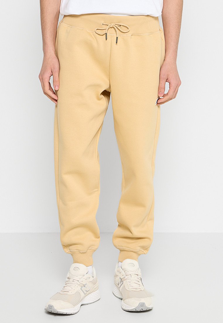 Pantalones de chándal beige con cintura elástica, cordón ajustable, bolsillos laterales y puños acanalados. Llevados con zapatillas deportivas de color claro.