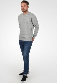 Pull en tricot gris clair avec texture côtelée, col rond et manches longues, associé à un jean bleu foncé et des baskets noires.