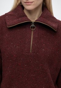 Femme portant un pull en maille bordeaux avec un grand col rabattu et une fermeture éclair dorée partiellement ouverte avec un curseur circulaire.