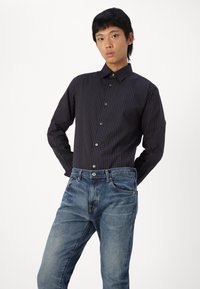 Camisa de rayas marinas de manga larga con cuello abotonado, combinada con unos vaqueros azules descoloridos. La tela tiene una textura suave; los botones son oscuros.