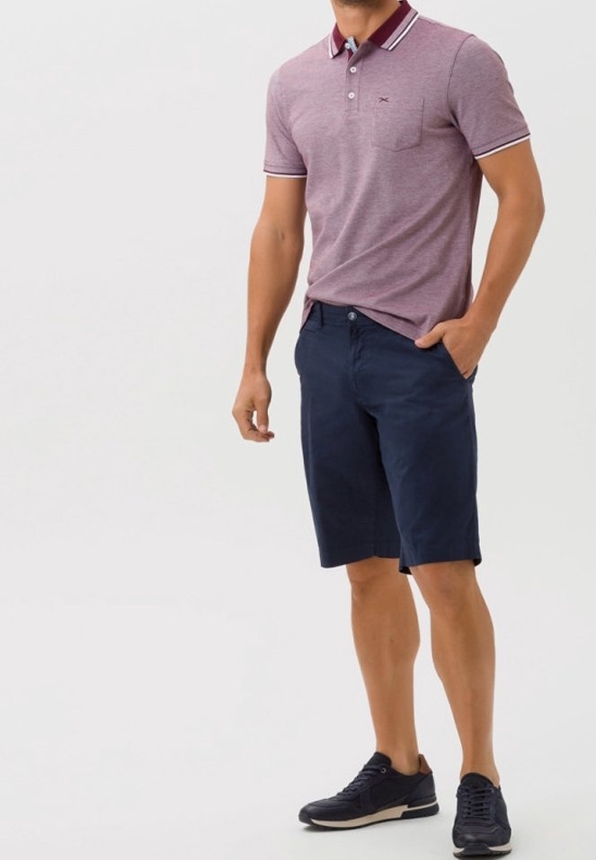 Homme portant un polo bordeaux à manches courtes, un short bleu marine et des baskets noires, debout avec une main dans la poche sur un fond uni.