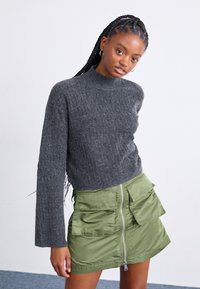 Pull long à manches longues gris à côtes avec un col montant, associé à une jupe mini zippée verte dotée de deux poches avant et d'une texture lisse.