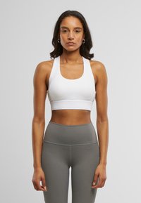 Hvit sports-BH med brede stropper og dyp u-formet hals, med en glatt tekstur og tettsittende design. Sammen med høyhælte grå leggings.