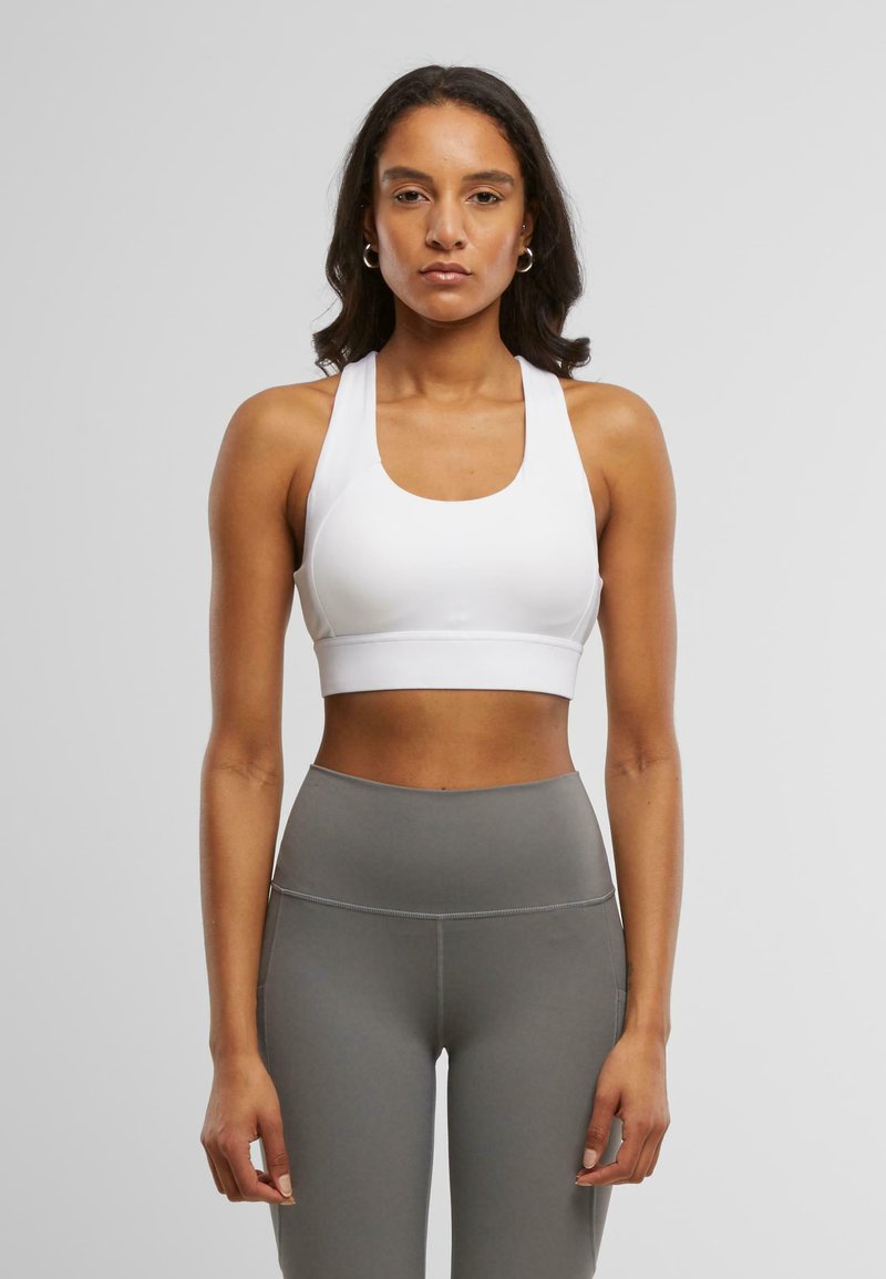 Hvit sports-BH med brede stropper og dyp u-formet hals, med en glatt tekstur og tettsittende design. Sammen med høyhælte grå leggings.