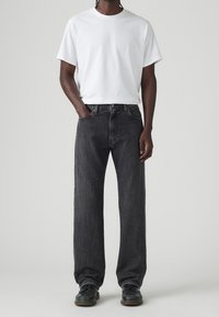 Muž stojící s rukama částečně v kapsách, oblečený v jednoduchém bílém tričku, volných černých džínách a černých botách na jednoduchém světlém pozadí.