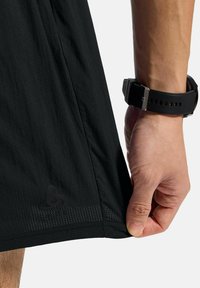 Zwarte sportshorts van een gestructureerde stof, met reflecterende details en een logo aan de zoom, vastgehouden door een hand met een zwarte horloge.