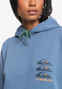 Mėlyna kapučinė džemperis su tekstūruotu audiniu, reguliuojama žalia virvelė ir juodas grafikos dizainas su kalnais bei „Quiksilver“ logotipu.