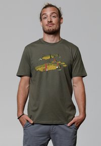 Olijfgroen katoenen t-shirt met een speelse afbeelding van kabouters, een ladder en kleurrijke elementen op een zandkleurige achtergrond.