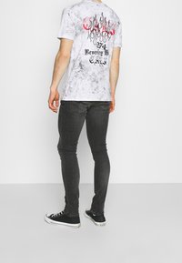 Bílé grafické tričko s červeno-černým motivem plamene na zádech, doplněné tmavě šedými úzkými džíny a černými teniskami.