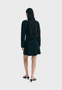 Schwarzes Kleid mit langen Ärmeln, tailliertem Oberteil und ausgestelltem Rock. Sanfte Textur und bis zur Mitte des Oberschenkels reichend. Getragen mit schwarzen High-Heels.