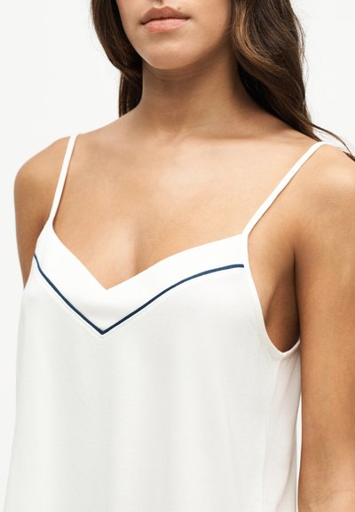 Femme portant un débardeur blanc avec de fines bretelles et une bordure bleu marine inclinée le long du décolleté, vue du cou jusqu'au milieu du torse.