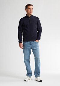 Granatowa bluza zapinana na zamek z teksturowanym wzorem, w połączeniu z jasnoniebieskimi dżinsami i białymi trampkami. Prosty design, swobodny krój i jednolite kolory.