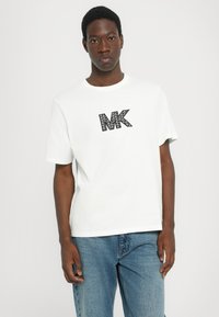Hvit t-skjorte med korte ermer som har en svart trykt "MK"-logo. Laget av bomull, med rund hals og avslappet passform. Uformelle denimjeans.