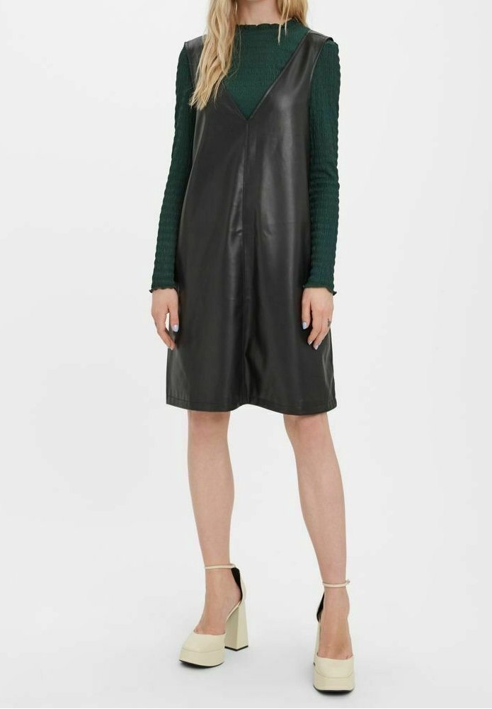 Femme portant une robe en cuir noire sans manches par-dessus un top vert à manches longues, assortie à des chaussures crème à talons hauts avec bride à la cheville.