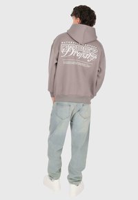 Osoba oblečená v malinovom mikinovom svetri s bielym potlačeným textom na chrbte, v kombinácii s svetlo modrými džínsy a bielymi teniskami, stojaca na neutrálnom pozadí.