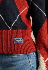 Punainen ja tummansininen neulepaita, jossa on timanttikuvioita ja valkoisia yksityiskohtia. Ribbattu kaulus ja hihansuut. Tunnuslappu lukee "Superdry Outdoor Outfitter."