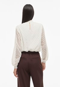 Weiße Bluse mit Chevron-Muster, mit hohem Kragen, Ballonärmel und einem Schlitz im Rücken. Kombiniert mit schokoladenbraunen Hosen.