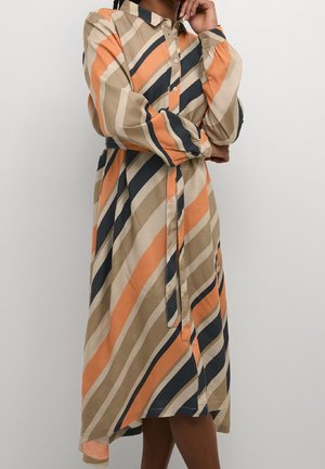 Femme portant une robe midi à manches longues boutonnée avec des rayures diagonales orange, beige et noires, debout devant un fond uni.