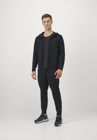 Schwarze Sportjacke mit Kapuze, kombiniert mit einem dunklen T-Shirt und schwarzen schmal geschnittenen Hosen. Verfügt über Taschen und Sneakers mit Gummisohle.