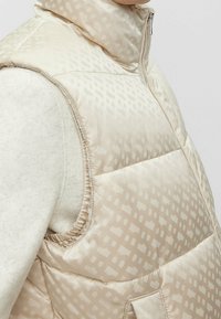 Beige puffer vest with a high collar, featuring a subtle pattern, zip closure, and side pockets. Smooth, slightly shiny texture.