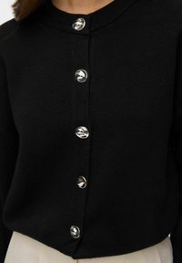 Cárdigan negro de punto con botones metálicos redondos y cuello redondo. Presenta una textura suave y un diseño recortado.