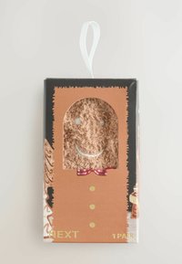 Braune Verpackung mit einem transparenten Fenster, das flauschige Socken mit einem Smiley-Gesicht und einer rot karierten Fliege zeigt, goldenen Text und dekorativen Rändern.