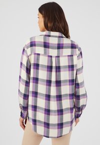 Camisa de franela de manga larga en color crema, con un patrón de cuadros en morado y gris, puños abotonados y un pliegue en la espalda para mayor facil de movimiento.