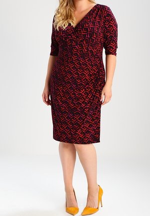 Mujer con un vestido hasta la rodilla de estampado rojo y negro con mangas tres cuartos y zapatos de tacón amarillo, de pie contra un fondo liso.