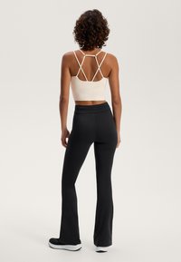 Világos bézs cropped top, keresztbe álló pántokkal, fekete, harangszárú nadrággal párosítva. Sima textúra, minimalista dizájn és sportos stílus.