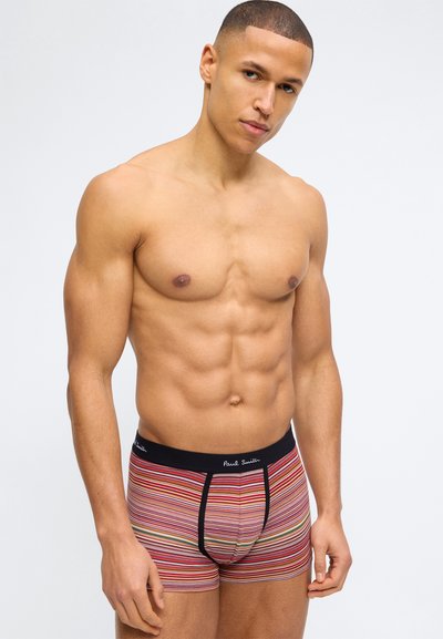 Homme musclé debout devant un fond uni, portant un boxer à rayures multicolores avec une ceinture noire et le logo Paul Smith.