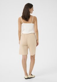 Beige lightweight shorts with an elastic waistband, paired with a white ribbed sleeveless top. Model facing away, showing full back view.