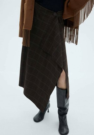 Asymetryczna ciemna spódnica w kratę z przednim rozcięciem, zestawiona z czarnymi wysokimi kozaczkami na obcasie i brązowym frędzlowanym płaszczem.