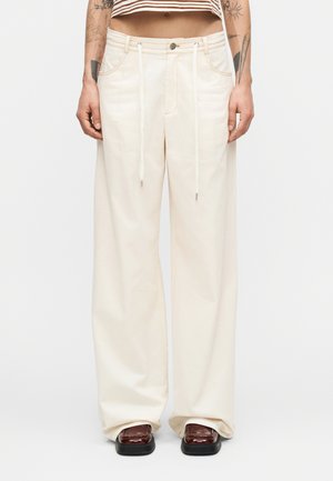 Άτομο που φορά χαλαρό εκρού παντελόνι με κορδόνια, κοντόριζη ριγέ μπλούζα και σκούρα καφέ παπούτσια με τετράγωνη μύτη, όρθιο μπροστά σε λευκό φόντο.