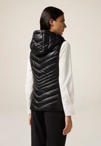 Schwarze gepolsterte Weste mit Kapuze, mit einem gestreiften Steppmuster und glänzender Oberfläche. Getragen über einem weißen Langarmshirt und schwarzen Hosen.