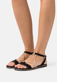 Black flat sandals featuring a suede finish, a gold accent strap design, and an adjustable ankle buckle. Open toe with minimalistic styling.