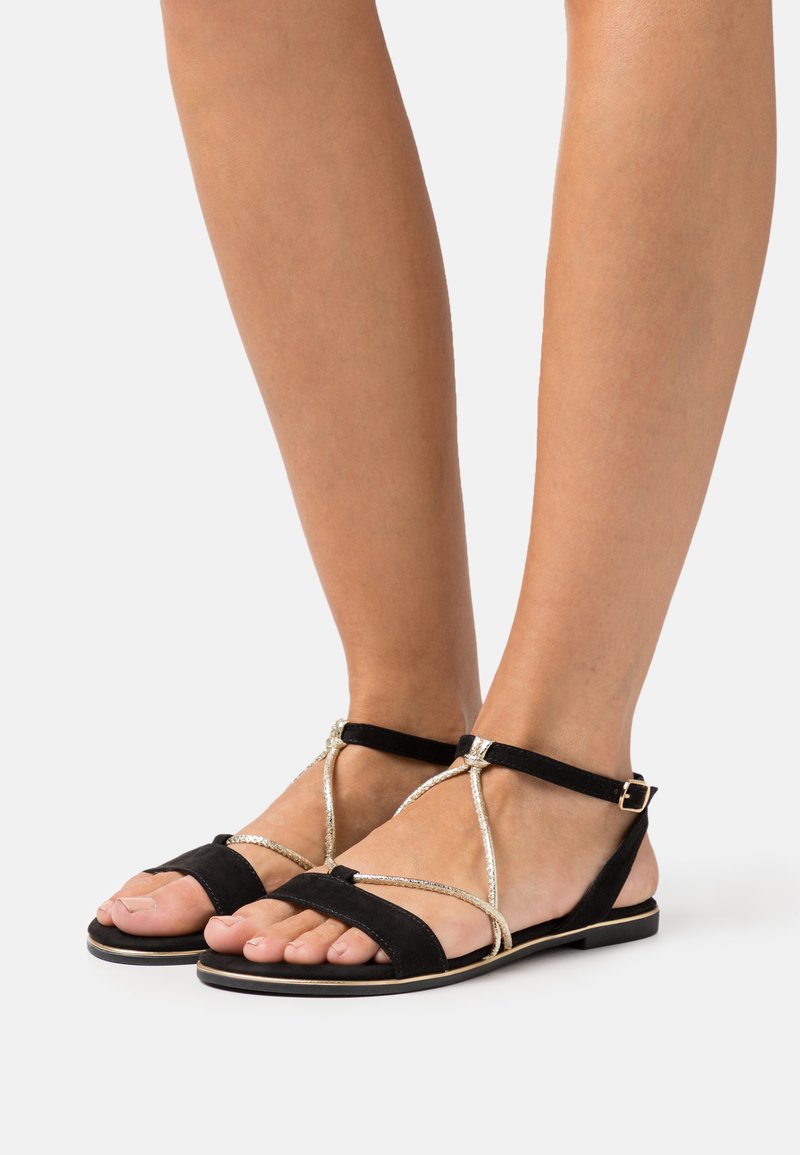 Black flat sandals featuring a suede finish, a gold accent strap design, and an adjustable ankle buckle. Open toe with minimalistic styling.