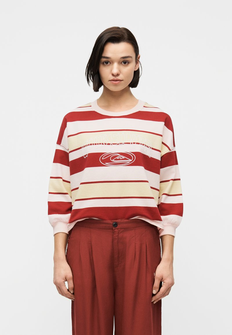 Νεαρή γυναίκα με κοντά σκοτεινά μαλλιά, που φοράει μακρυμάνικη ριγωτή μπλούζα σε κόκκινες, εκρού και μπεζ αποχρώσεις και παντελόνι ψηλόμεσο σε σκουροκόκκινη απόχρωση, στέκεται μπροστά σε λευκό φόντο.