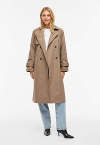 Trench-coat beige à double boutonnage avec de larges revers, manches longues et fentes latérales, associé à un jean droit bleu clair et des bottes noires à bout pointu.