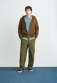 Καφέ φούτερ με φερμουάρ πάνω από ένα ανοιχτό γαλάζιο τζιν πουκάμισο, συνδυασμένο με φαρδιά χακί παντελόνια cargo και μαύρα αθλητικά παπούτσια σε μπλε μοκέτα.