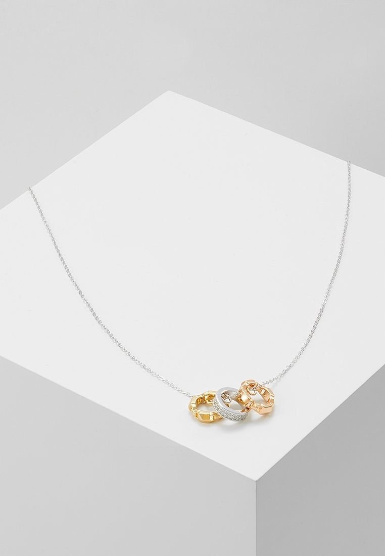 Collier délicat en chaîne d'argent avec trois anneaux reliés en or, argent avec diamants et or rose, présenté sur une surface géométrique blanche.