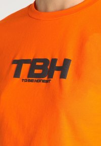 Camisa naranja brillante con grandes letras negras "TBH" y un texto más pequeño "TO BE HONEST" impreso en el pecho.