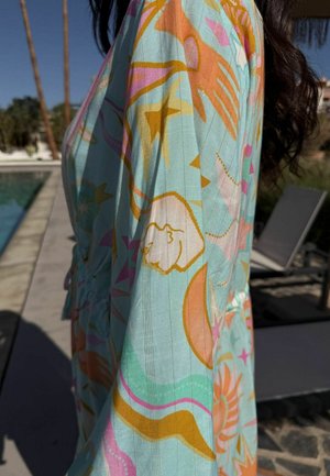 Nainen, joka pukeutunut värikkääseen, kuvioituun pitkähihaiseen vaatteeseen, seisoo uima-altaan vieressä, taustalla aurinkotuoleja ja palmuja.