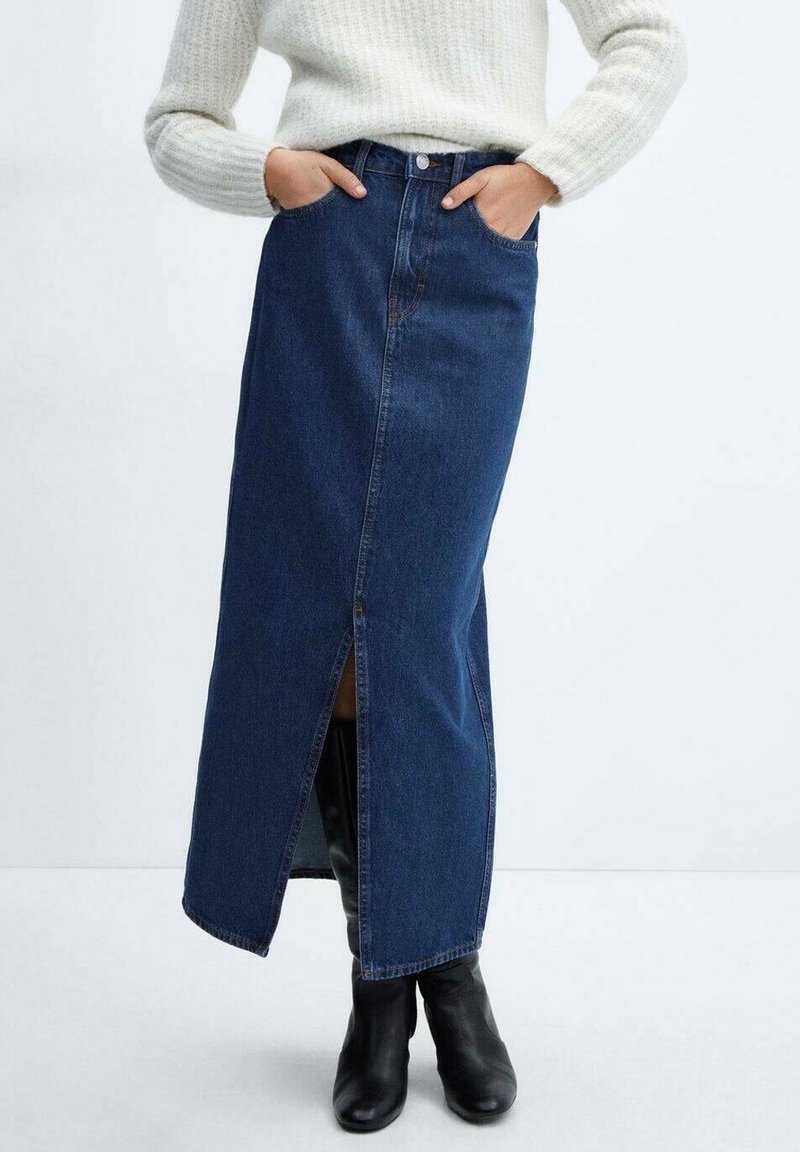 Femme portant un pull blanc en maille, une jupe midi en denim bleu foncé taille haute avec une fente à l'avant, et des bottes en cuir noir, les mains dans les poches.