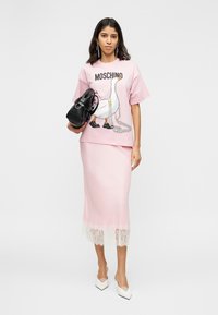 Femme portant un t-shirt rose Moschino avec un motif d'oie et un collier de perles, jupe assortie avec un ourlet en dentelle, tenant un sac noir, talons blancs.