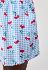 Blauwe ruitenrok met een kersenprint in het rood en groene bladeren. De stof lijkt lichtgewicht met een geplooide taille.