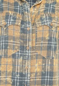 Camicia a quadri button-up nei toni del blu, giallo e grigio. Presenta due tasche sul petto, bottoni in legno e una texture leggermente invecchiata.