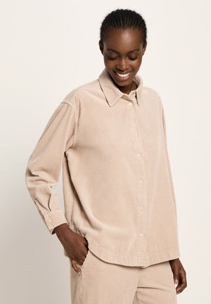 Smiling woman with braided hair wearing a beige corduroy button-up shirt and matching pants, one hand in pocket, looking down.
