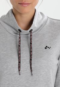 Sweat à capuche gris avec un col montant, doté de cordons noirs avec un texte rose. Petit logo noir sur la poitrine ; matière en coton doux.